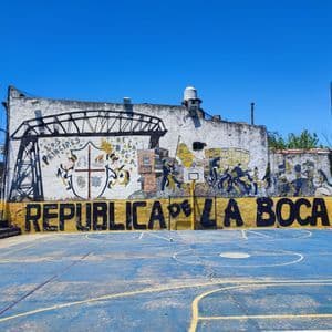 Un campo da basket blu vuoto davanti a un muro con un grande murale colorato che dice 'Republica de La Boca'.
