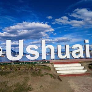 Il grande cartello bianco 'Ushuaia' si erge di fronte a un porto con barche sotto un cielo blu parzialmente nuvoloso.