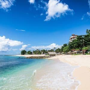 Playa tropical de arena con agua turquesa, palmeras y cabañas en la orilla bajo un cielo azul brillante con nubes blancas.