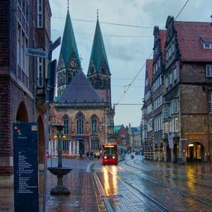 Un tranvía rojo con sus luces encendidas transita por una calle europea mojada al anochecer, con una catedral de dos torres en el fondo.