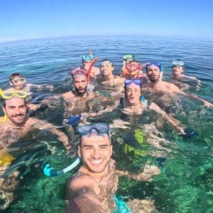 Un groupe WeRoad en voyage, équipé de masques et tubas, sourit pour un selfie en nageant dans une eau turquoise cristalline.