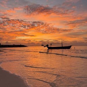 Ein silhouettiertes Boot schwimmt auf dem Meer nahe einem Sandstrand unter einem leuchtend orangefarbenen Himmel bei Sonnenuntergang.