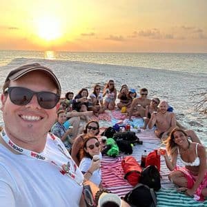 Eine WeRoad-Gruppe macht ein Selfie während eines Picknicks an einem Sandstrand bei Sonnenuntergang.