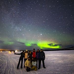 Un gruppo WeRoad in viaggio posa su una piana innevata di notte, con l'aurora boreale e le stelle visibili nel cielo.