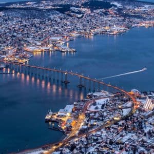 Una vista aerea di una città costiera innevata al tramonto, con un lungo ponte illuminato che attraversa l'acqua.