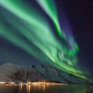 L'aurora boreale verde illumina il cielo notturno stellato sopra montagne innevate e un porto illuminato con barche.