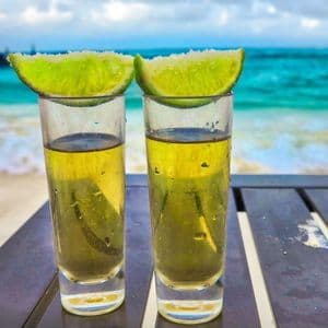 Due shot di tequila con spicchi di lime salati poggiano su un tavolo con vista su un oceano turchese e una spiaggia sabbiosa.