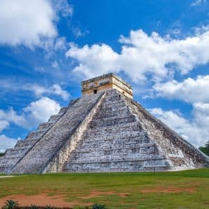 Una grande piramide Maya a gradoni su un campo erboso sotto un cielo blu parzialmente nuvoloso