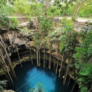 Una vista dall'alto di un profondo cenote pieno di acqua blu, con lunghe liane che pendono dalle scogliere rocciose circostanti e dalla giungla.