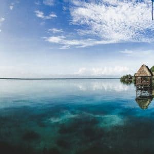 Bungalow dal tetto di paglia sulla riva di una laguna tranquilla si riflettono nell'acqua calma e turchese sotto un cielo blu.