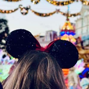 La nuca di una persona che indossa orecchie da topo nere con paillettes, con una parata sfocata e festosa sullo sfondo.