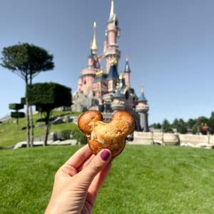 Una mano tiene un dolce a forma di Topolino di fronte a un grande castello da fiaba rosa su un prato verde.