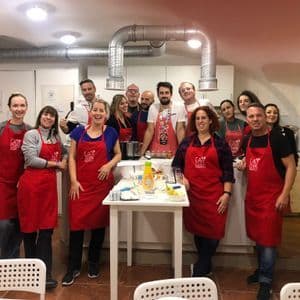 Un gruppo WeRoad sorride per una foto durante una lezione di cucina, tutti indossano grembiuli rossi coordinati in una cucina.