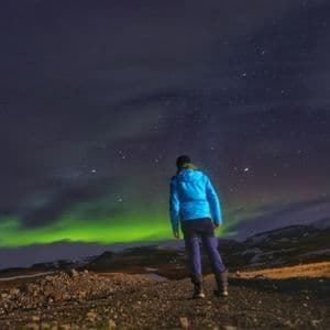 Una persona in giacca blu si trova su un sentiero di notte, osservando l'aurora boreale verde sotto un cielo stellato.