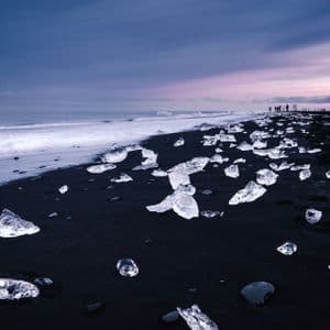 Un viaggio di gruppo WeRoad su una spiaggia di sabbia nera dove blocchi di ghiaccio sono sparsi dalle onde dell'oceano sotto un cielo crepuscolare.