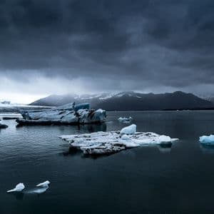 Iceberg galleggiano in una laguna glaciale di fronte a montagne innevate sotto un cielo scuro e nuvoloso, con due uccelli che riposano sul ghiaccio in primo piano.