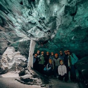 Un gruppo WeRoad con caschi e lampade frontali posa per una foto di gruppo all'interno di una grande grotta di ghiaccio con pareti turchesi.