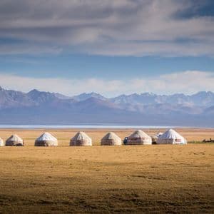 Una fila di yurte su una pianura erbosa di fronte a un lago e una catena montuosa distante.