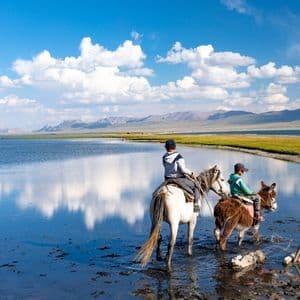 Deux personnes à cheval et à dos d'âne traversent un lac paisible reflétant les nuages et les montagnes lointaines.