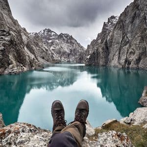 A first-person view of hiking boots on a rocky ledge, overlooking a calm turquoise lake surrounded by steep mountains under a cloudy sky.