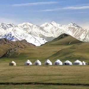 A row of yurts sits on a wide green plain, with rolling hills and a vast, snow-capped mountain range behind them.