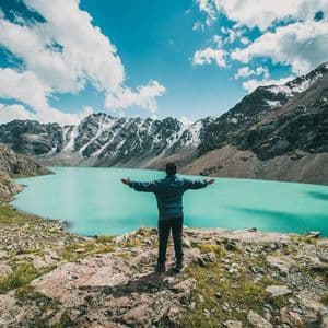 Eine Person steht mit ausgestreckten Armen auf einem Felsvorsprung und blickt über einen türkisen Alpensee, der von schneebedeckten Bergen umgeben ist.