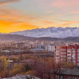 Un paysage urbain avec des bâtiments nichés devant une chaîne de montagnes enneigées sous un ciel de coucher de soleil doré.