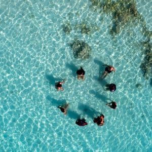 Vista aerea di un gruppo WeRoad in cerchio in acqua bassa, limpida e turchese.