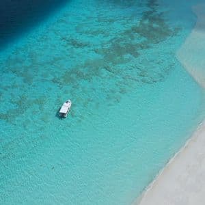 Una vista aerea di una barca bianca in acqua turchese accanto a una lingua di sabbia bianca, in contrasto con il mare blu profondo.