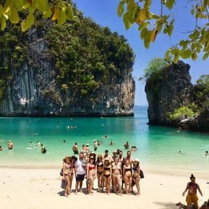 Un grupo de WeRoad posa en una playa de arena en una bahía turquesa, con grandes acantilados cubiertos de árboles al fondo.