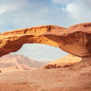 Un arco naturale di arenaria incornicia montagne lontane in un paesaggio desertico sotto un cielo parzialmente nuvoloso.