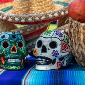 Dos calaveras de azúcar coloridas reposan sobre un serape a rayas junto a una cesta de frutas y sombreros.