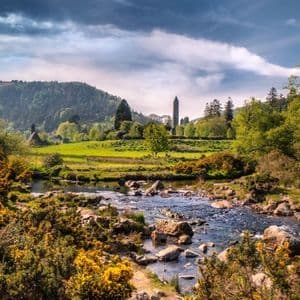 Un ruscello roccioso scorre attraverso una valle verde e rigogliosa con fiori gialli, con una torre in pietra e una collina boscosa sullo sfondo.