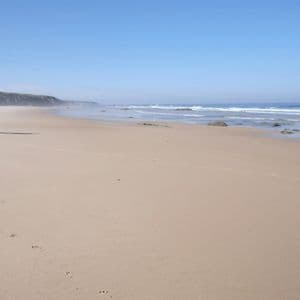 Una persona con un grande zaino cammina a piedi nudi su un'ampia spiaggia sabbiosa, lasciandosi dietro delle impronte.
