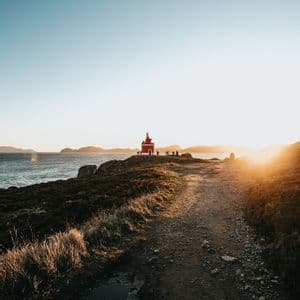 Un gruppo WeRoad si trova accanto a un piccolo faro rosso in cima a una scogliera costiera al tramonto, con un sentiero sterrato in primo piano.