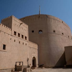 Un imponente forte color sabbia con una torre rotonda domina un cortile in pietra contenente un pozzo e cannoni, sotto un cielo blu limpido.