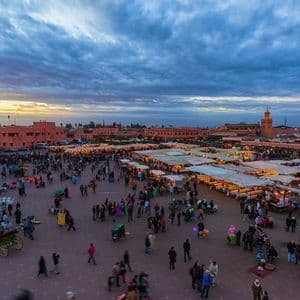 An elevated view of a bustling city square crowded with people and market stalls at dusk, with terracotta buildings in the background.