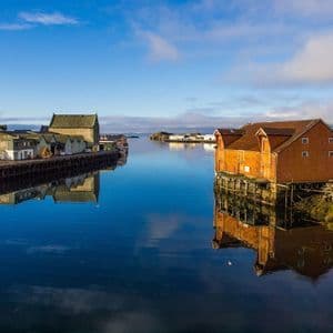 Casas de botes de madera sobre pilotes bordean una ensenada tranquila, sus reflejos espejados en el agua azul inmóvil bajo un cielo despejado.