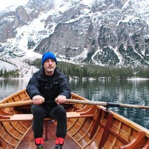 Un uomo con un berretto blu rema una barca di legno su un lago con montagne innevate sullo sfondo.