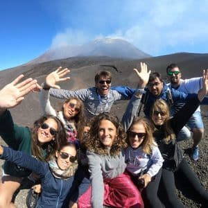 Un grupo de WeRoad se toma un selfi en una ladera rocosa, con un volcán humeante al fondo bajo un cielo azul claro.