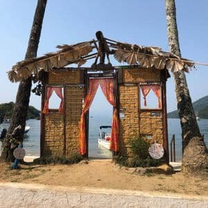 A rustic mud-brick gateway with a thatched roof stands between two palm trees on a beach, framing a view of the sea with a boat.