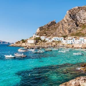Small boats anchored in a turquoise bay in front of a coastal village with white houses built into the side of a rocky mountain.