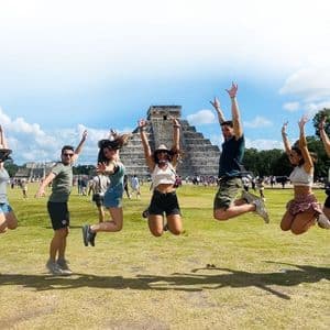 A WeRoad group trip jumping in the air on a grassy lawn in front of an ancient stone pyramid under a sunny sky.
