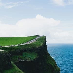 Un viaggio di gruppo WeRoad percorre un sentiero in cima a ripide scogliere verdi a picco sull'oceano, sotto un cielo nuvoloso.
