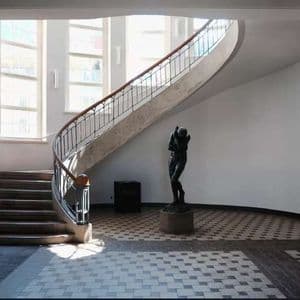 Una sala illuminata dal sole con una scala curva in cemento, una scultura scura su un piedistallo e un pavimento a scacchi.