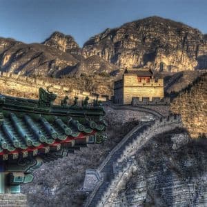 Una larga muralla de piedra con una torre de vigilancia serpentea a lo largo de una montaña escarpada, con la esquina de un tejado de tejas verdes en primer plano.