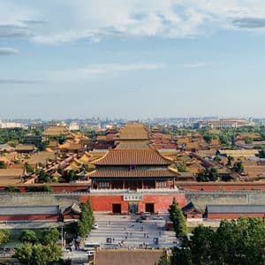 Una vista elevada de un vasto complejo palaciego asiático tradicional con tejados de tejas naranjas, con un paisaje urbano moderno visible al fondo.