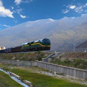 Un tren de carga verde y amarillo viaja por las vías a través de un vasto paisaje montañoso bajo un cielo parcialmente nublado.