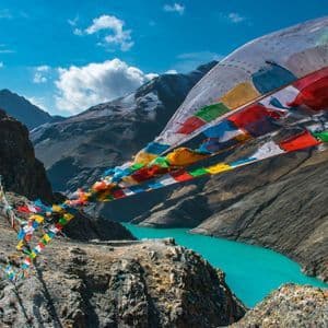 Coloridas banderas de oración ondean al viento sobre un río turquesa que serpentea por un valle de escarpadas montañas nevadas.