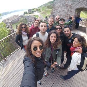 Un groupe WeRoad prend un selfie souriant sur une passerelle en bois avec des ruines de château et un plan d'eau en arrière-plan.
